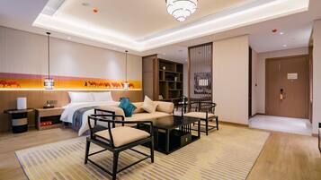 Een hotelkamer met een bed, een bank, stoelen, een salontafel, een nachtkastje, een lamp, een boekenplank en een deur.