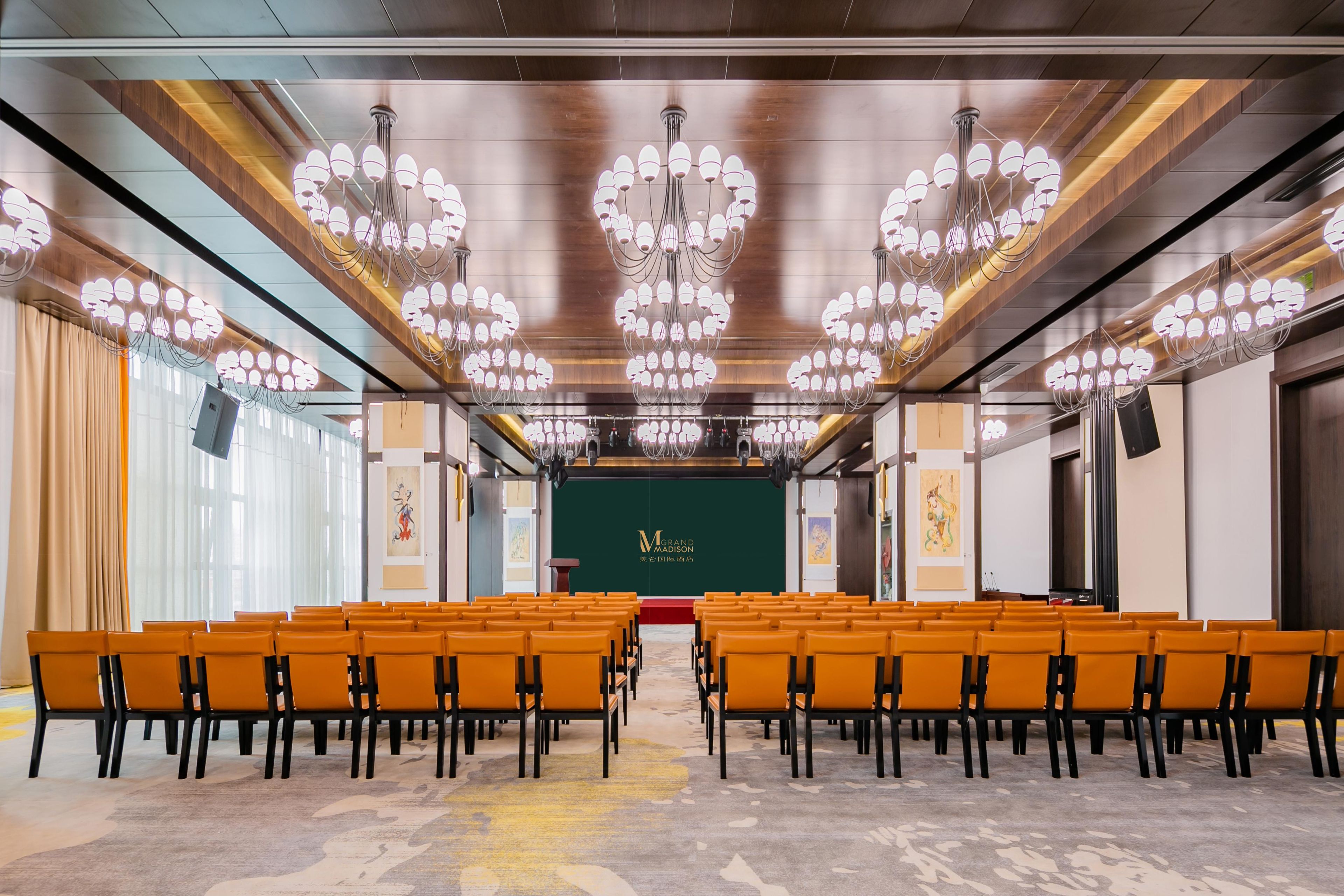 A conference room with rows of orange chairs, a stage with a green backdrop, and multiple chandeliers.