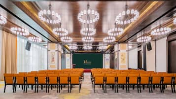 A conference room with rows of orange chairs, a stage with a green backdrop, and multiple chandeliers.
