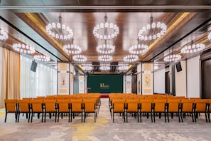 A conference room with rows of orange chairs, a stage with a green backdrop, and multiple chandeliers.