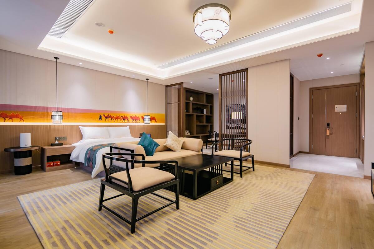 Ein Hotelzimmer mit Bett, Sofa, Sesseln, Couchtisch, Nachttisch, Lampe, Bücherregal und einer Tür.