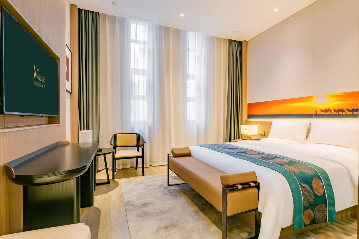 Ein Hotelzimmer mit Bett, Schreibtisch, Stuhl, Bank, Fernseher und einem Sonnenuntergangsbild.