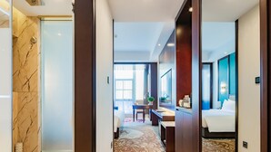 A hotel corridor with a marble floor, a bed with a blue headboard, and a desk with a plant. - Grand Madison Xuchang Beihai (Xuchang)