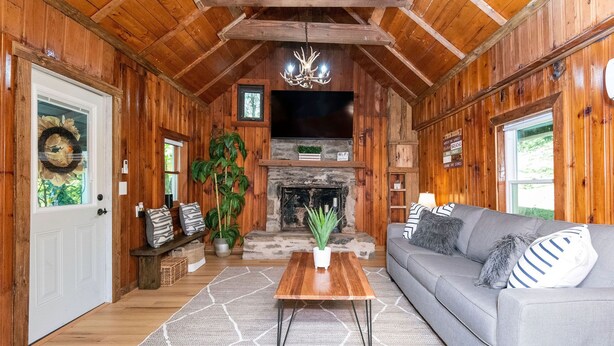 A cozy cabin interior with a fireplace, wooden walls, a grey sofa, and a wooden coffee table. - Cozy Mountain Cabin w/ hot tub and fire pit (Boone)