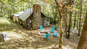 A rustic cabin with a stone chimney, surrounded by a forest. There are four turquoise Adirondack chairs arranged around a fire pit. - Cozy Mountain Cabin w/ hot tub and fire pit (Boone)