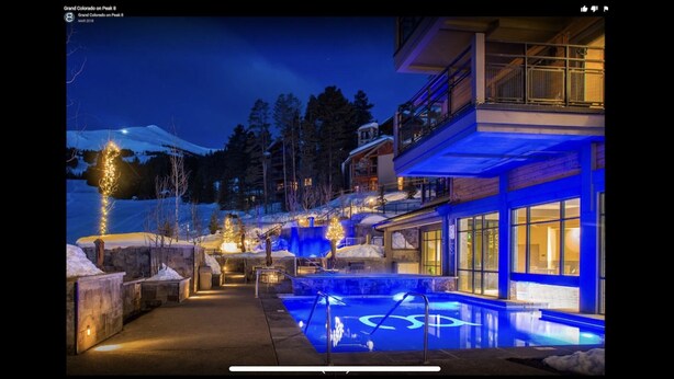 A modern hotel with a pool area, illuminated by blue lighting, surrounded by snow-covered landscape and trees.