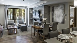 A modern hotel room with a dining area, a sofa set, a flat-screen TV, and a large abstract painting.