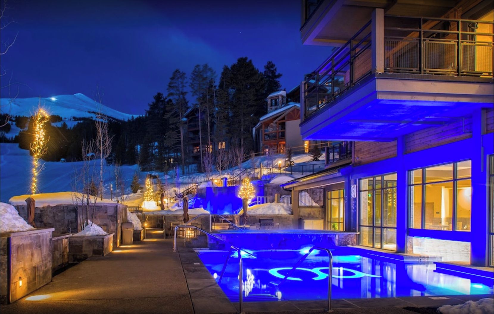 Un hotel moderno con zona de piscina, iluminada con luces azules, rodeado de paisaje nevado y árboles.