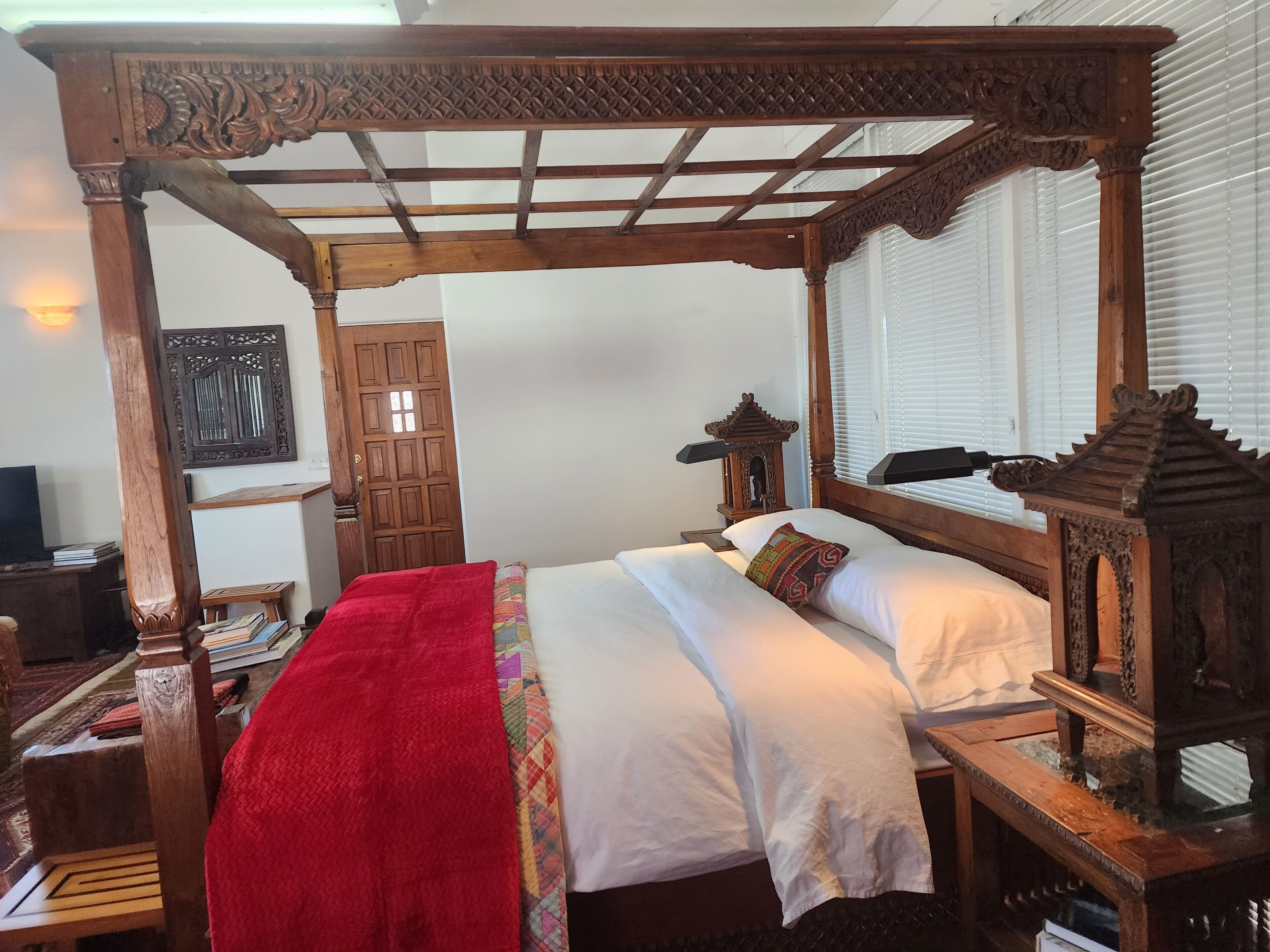 Een traditioneel houten hemelbed met een rode deken en kussens, een nachtkastje van hout met een lamp, en een kleine houten constructie die op een lantaarn lijkt.