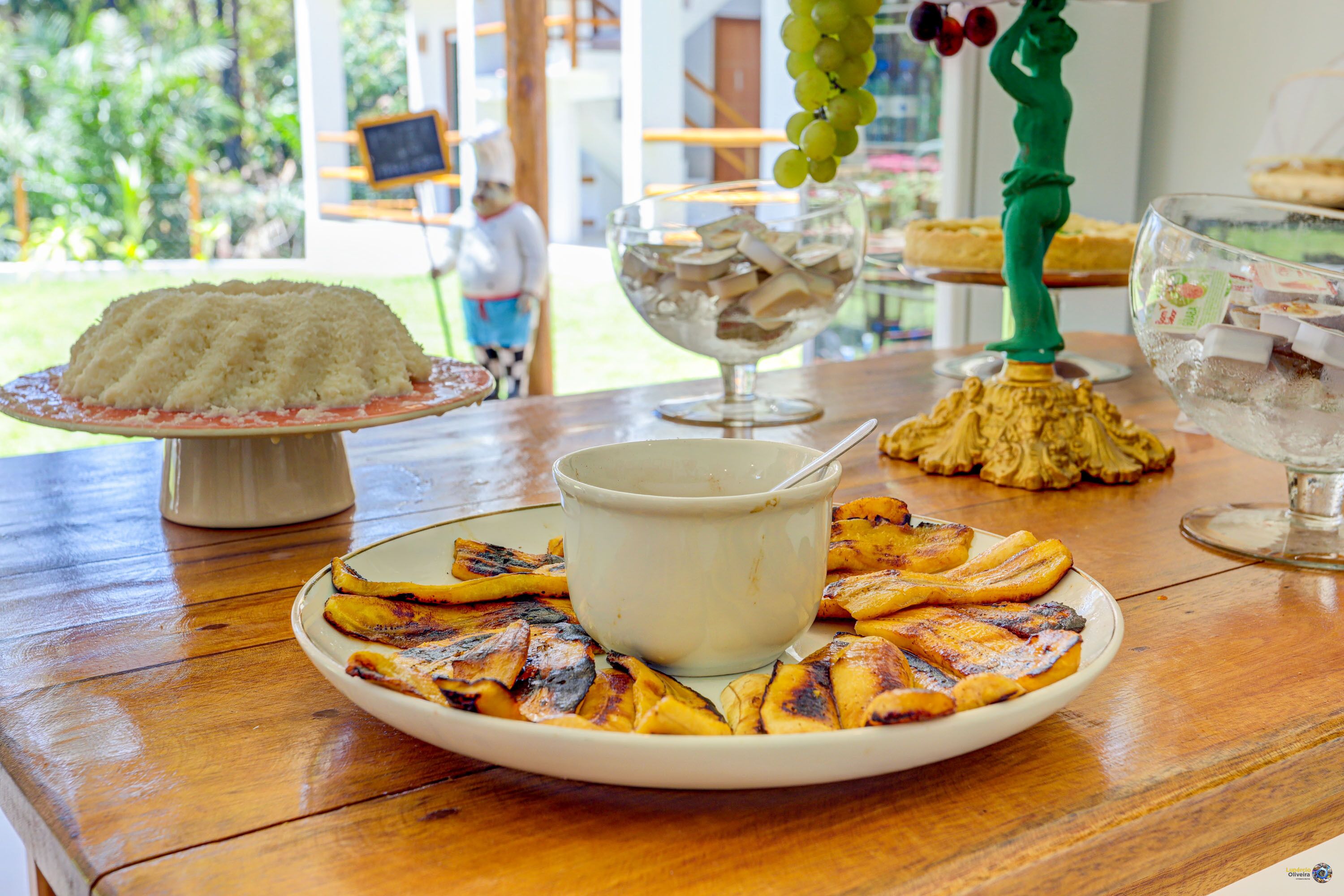 A wooden table with a plate of grilled bananas, a bowl of white sauce, and a decorative vase with grapes.