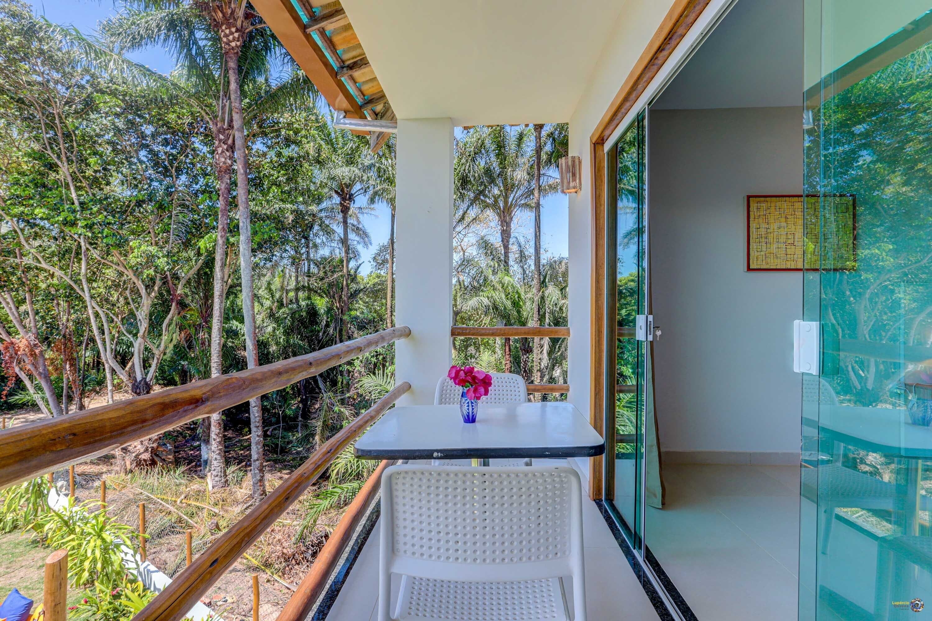 A balcony with a glass door, a white chair, a small table with a vase of flowers, and a view of dense tropical foliage.