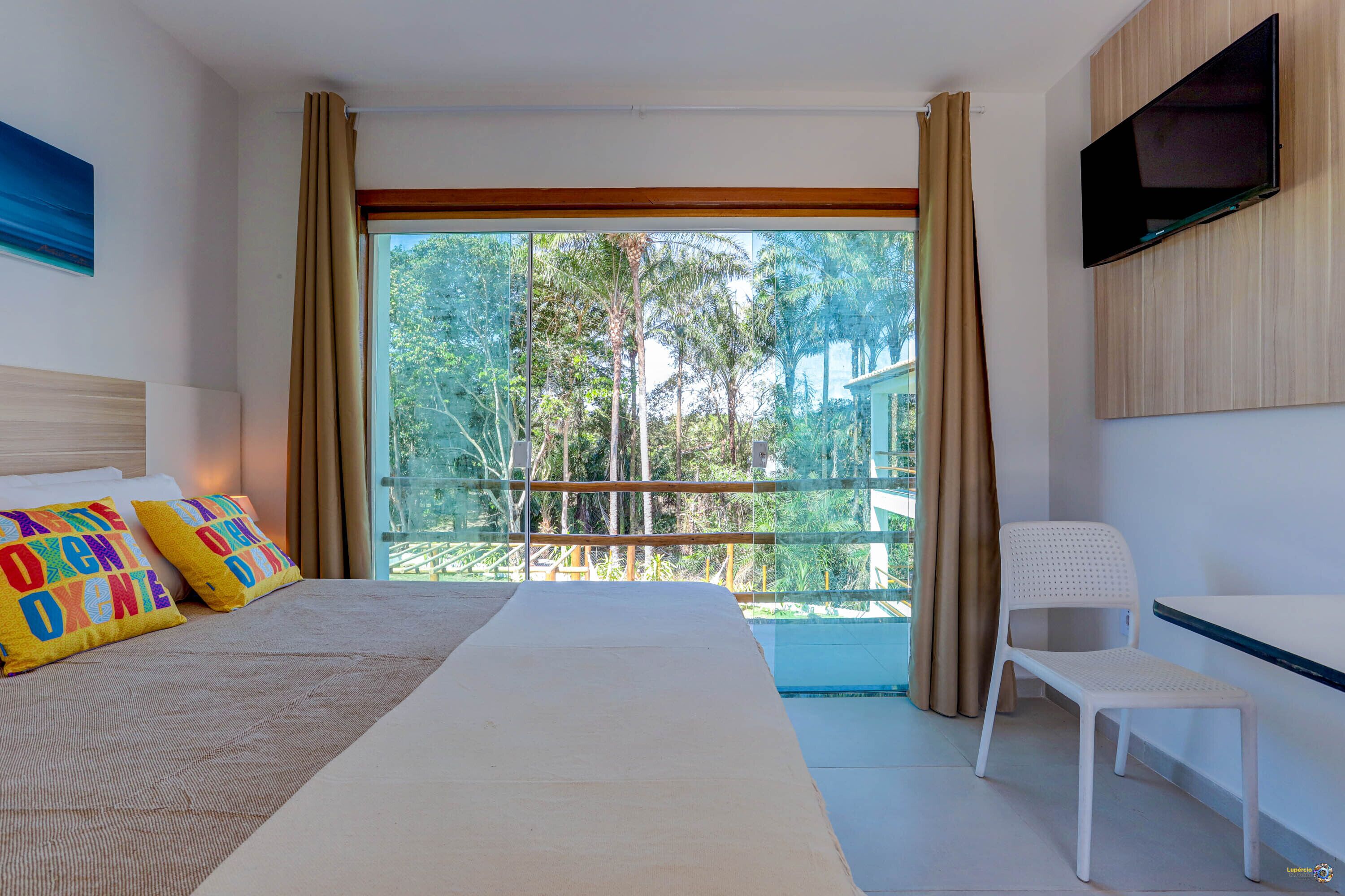 A hotel room with a bed, a chair, a TV, and a balcony with a view of trees.