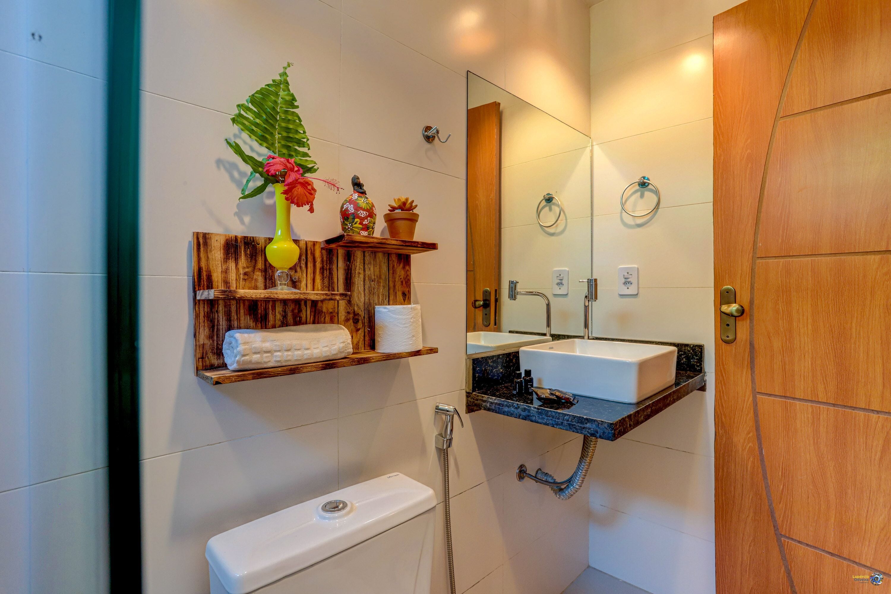 Un baño con estante de madera con artículos de aseo, inodoro, lavamanos con espejo y ducha.