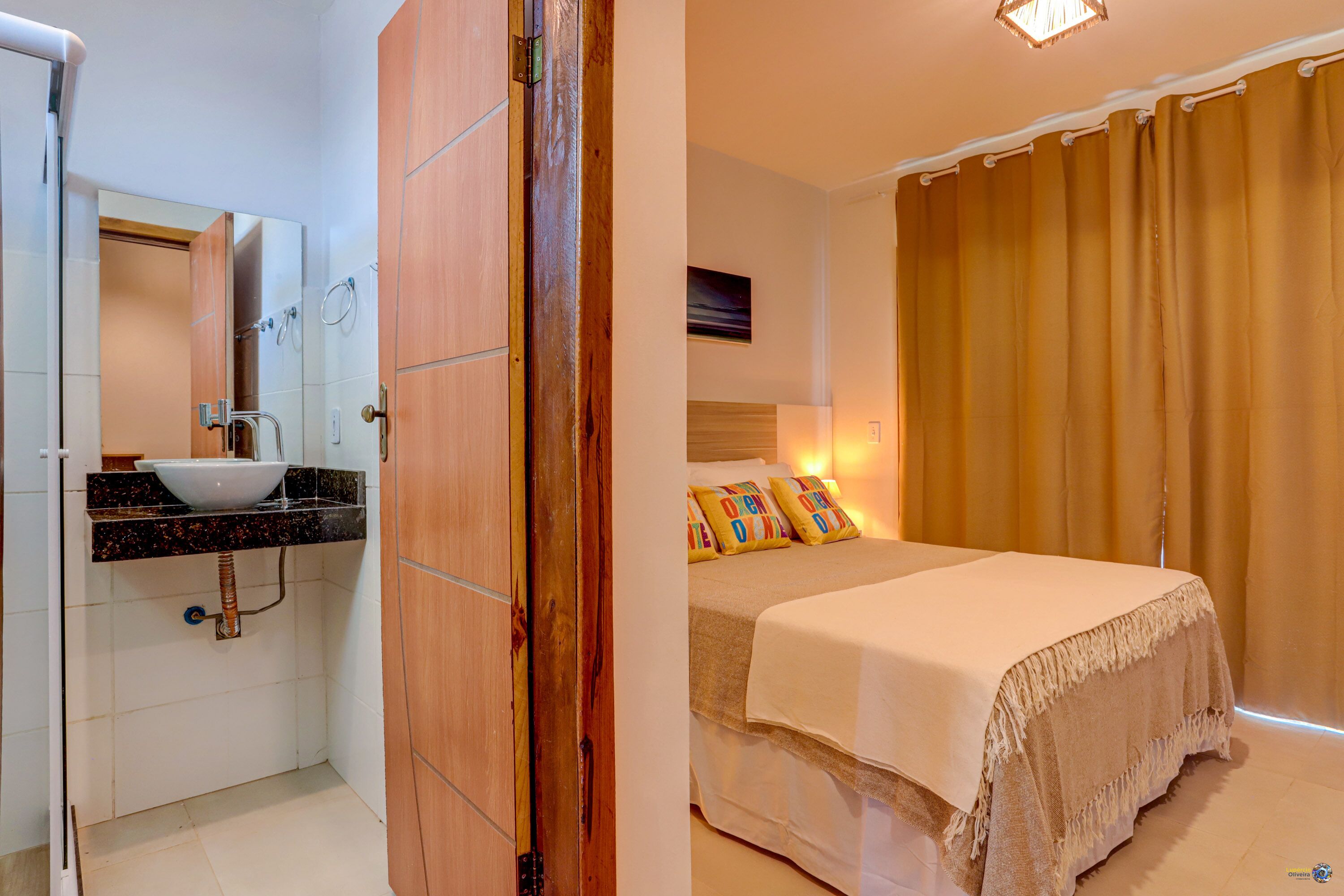 Baño con lavamanos, espejo y ducha. La habitación tiene una cama con colcha y almohadas blancas.
