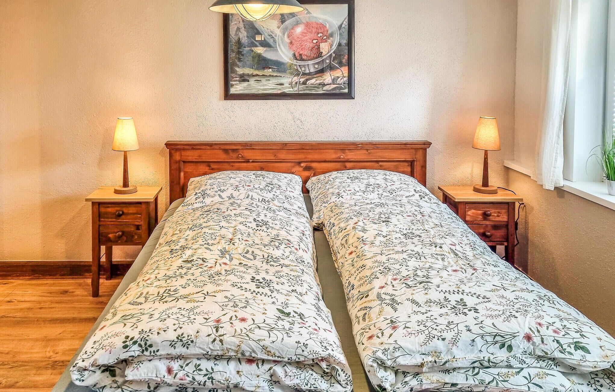 Een tweepersoonsbed met bloemenbeddengoed, twee nachtkastjes met lampen, een inlijstte afbeelding aan de muur en een raam met gordijnen.
