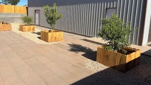A building with a corrugated metal exterior, surrounded by a paved area with wooden planters containing small trees. - Quiet, Cozy Three Bedroom Two Bath Home, Great Location (Page)