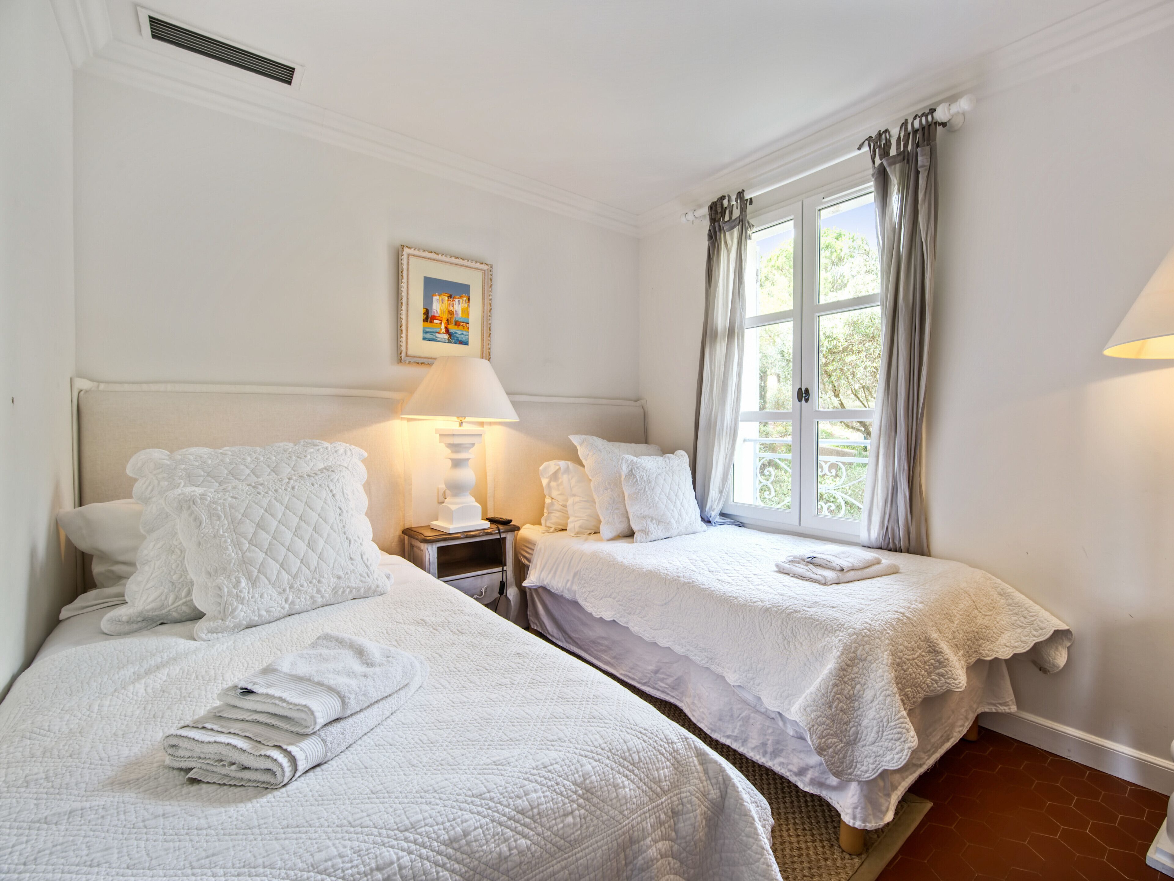 Quarto com duas camas, uma mesinha de cabeceira com abajur, um quadro na parede e uma janela com cortinas.