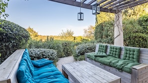 Eine hölzerne Pergola mit einer Bank und einem Tisch, umgeben von üppigem Grün und blauem Himmel.