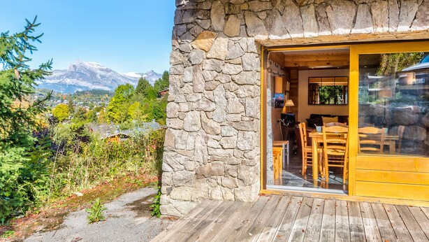 Pared exterior de piedra, terraza de madera y zona de comedor con sillas y una mesa.