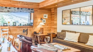 Interior acogedor de una cabaña con una mesa de madera, sillas de cuero, un sofá y una ventana con vistas a un paisaje nevado.