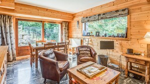 Interior acogedor de una cabaña con comedor, sillones de cuero, una mesa de centro con libros, televisor y ventana con vistas a follaje.