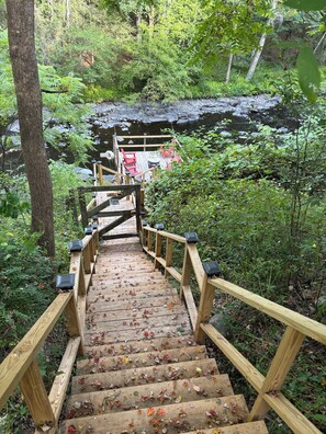 A wooden staircase leading to a wooden deck with red chairs and a table, surrounded by dense greenery and a rocky stream. - Rustic two bedroom cottage on riverside in Shohola PA (Shohola)