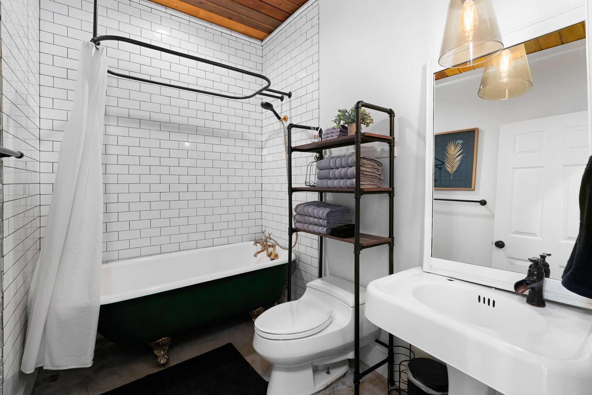 Et badeværelse med et hvidt badekar, toilet, hvid vask og et spejl. Der er en håndklædestang med sammenfoldede håndklæder og en hylde med en plante.
