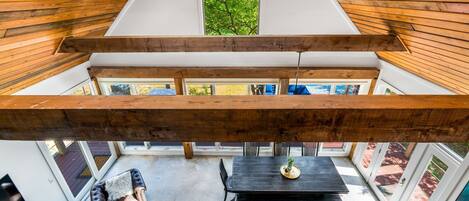 A loft-style room with a high ceiling, exposed wooden beams, a central skylight, a dining area with a table and chairs, and a view of the outdoors.