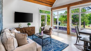 A spacious living room with a large TV, comfortable seating, and a view of the outdoors through large windows. - Elegant Birdhouse Cabin with 🔥Heated🔥 Cowboy Pool (Kyle)