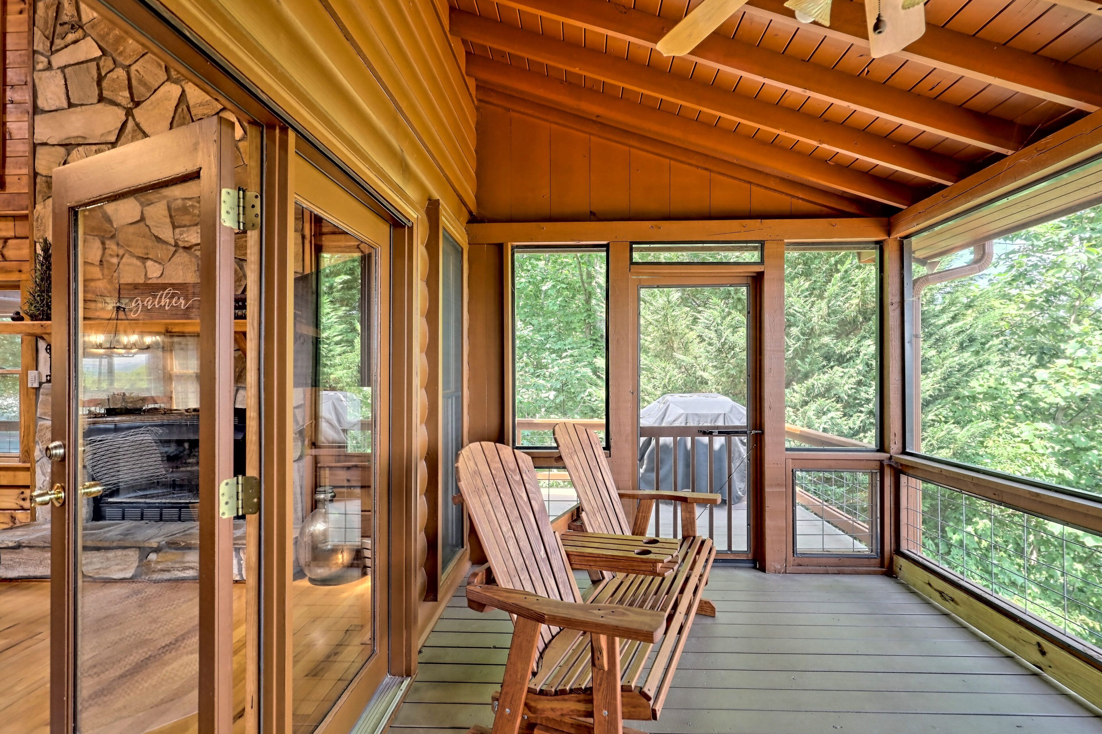 A wooden deck with Adirondack chairs, a stone fireplace, and large windows offering a view of the outdoors.
