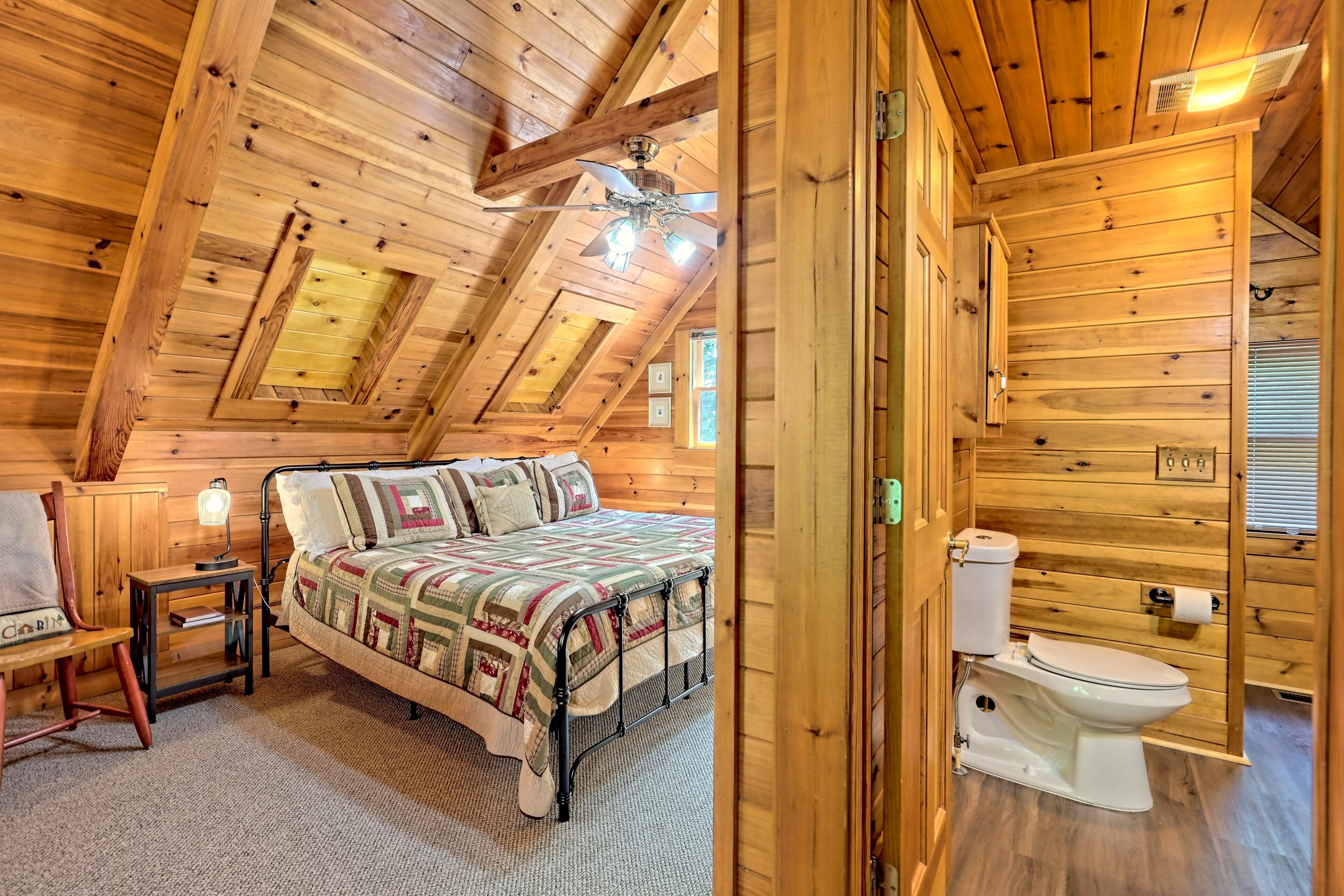 Ein Schlafzimmer in einer Holzhütte mit Bett, Stuhl, Nachttisch, Deckenventilator und Toilette.