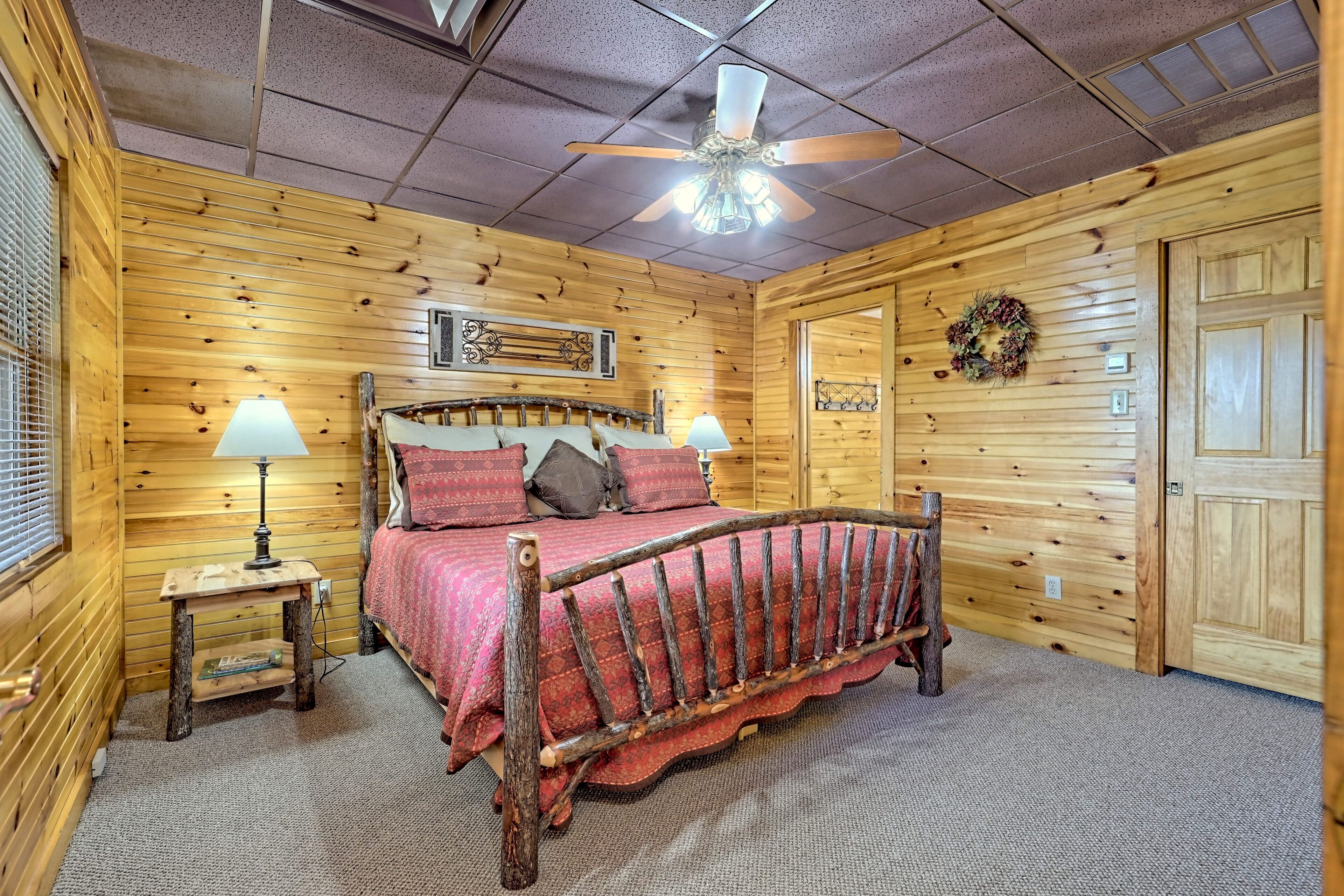 A wooden-paneled bedroom with a bed, two bedside lamps, a ceiling fan, and a door with a wreath.