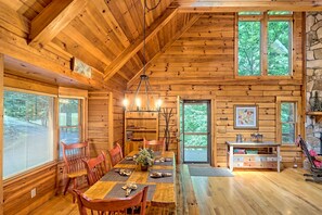 A wooden cabin interior with a dining table, chairs, a chandelier, and large windows.