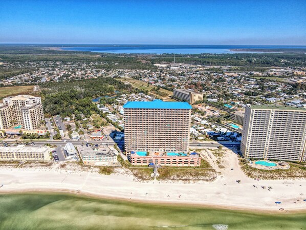 A high-rise hotel with a blue roof, located near a beach with a pool and a parking area.