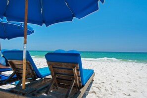 A beach scene with two wooden lounge chairs under blue umbrellas, facing the ocean.