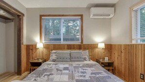 Un dormitorio con pared de paneles de madera, una cama con ropa de cama estampada, dos lámparas de noche y una ventana con persianas.