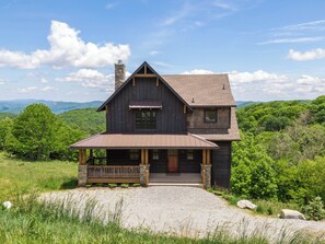 A two-story log cabin with a stone chimney, a porch with a bench, and a gravel driveway.