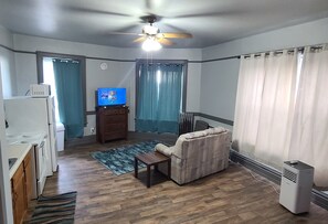 A living room with a sofa, a television, a ceiling fan, and a kitchen area with a microwave.