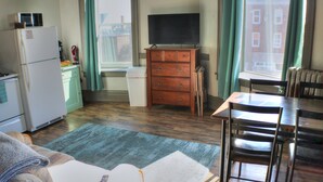 A compact kitchen with a refrigerator, a microwave, and a small oven. There is a wooden cabinet with a television on top. A dining table with chairs is positioned near a window with curtains.