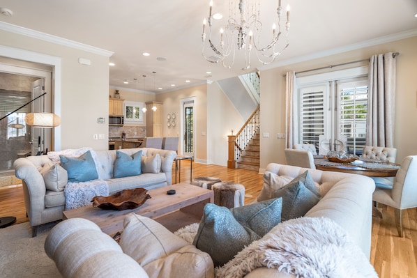 A spacious living room with a large sofa, a coffee table, and a chandelier. There is a staircase leading to an upper level, and a kitchen area is visible in the background.