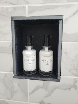 Two hand-crafted conditioner and shampoo bottles in a black-framed shelf against a white marble wall.