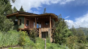 A rustic wooden house with a balcony, surrounded by lush greenery and a clear sky.
