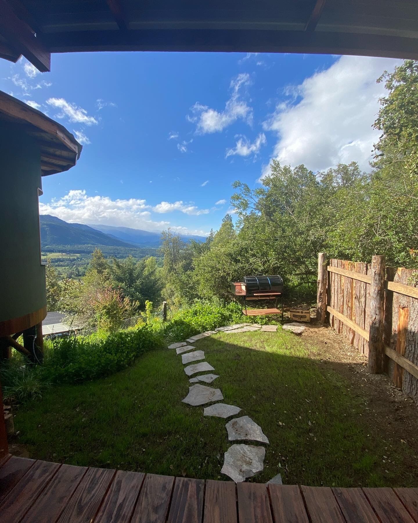 Eine Holzterrasse führt zu einem Steinweg in einem üppigen Garten mit Blick auf ferne Berge und einen klaren Himmel.