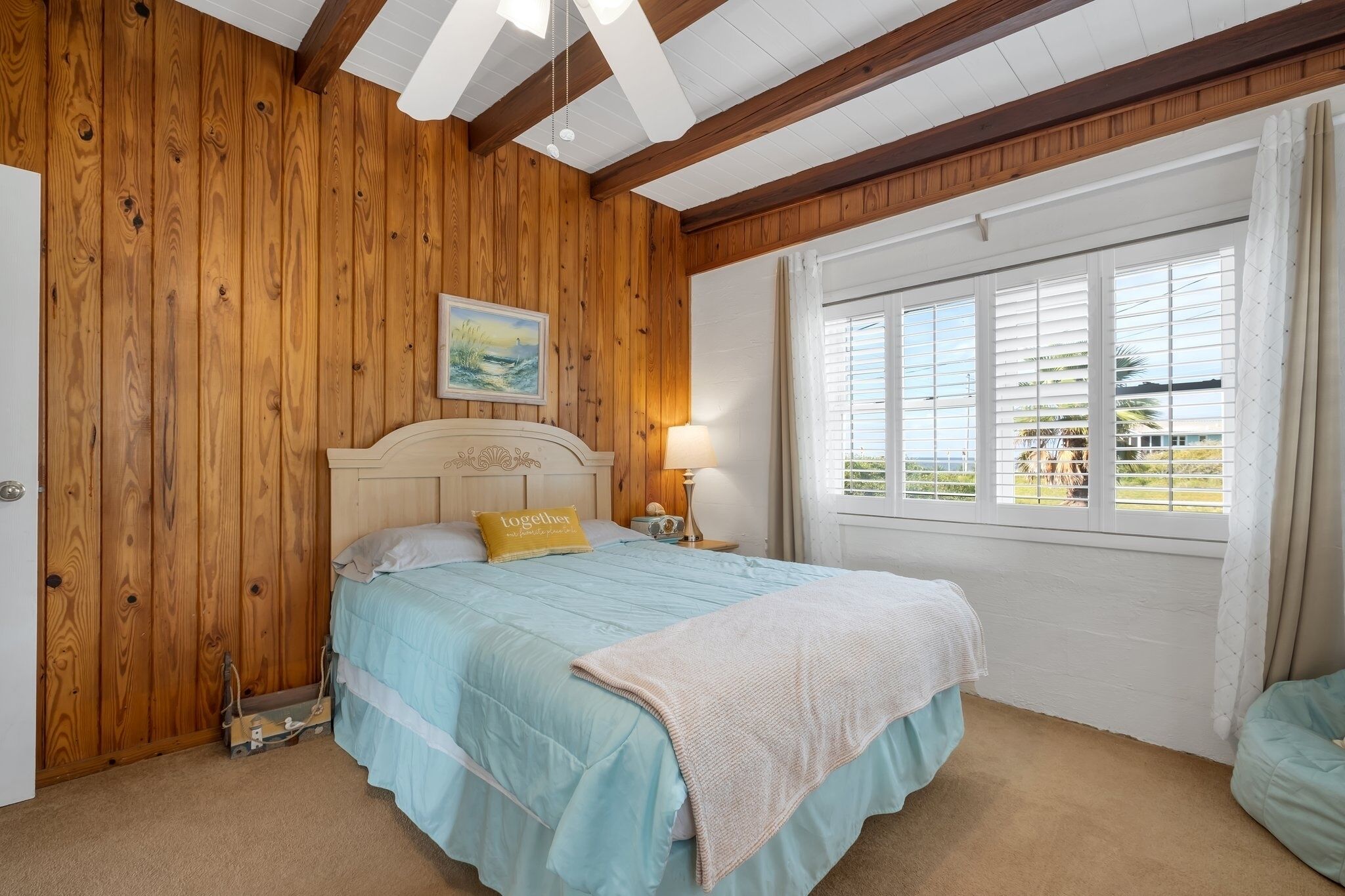 A bedroom with a wooden wall, a bed with a blue bedspread, a yellow pillow, a nightstand with a lamp, and a window with blinds.