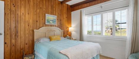 A bedroom with a wooden wall, a bed with a blue bedspread, a yellow pillow, a nightstand with a lamp, and a window with blinds.