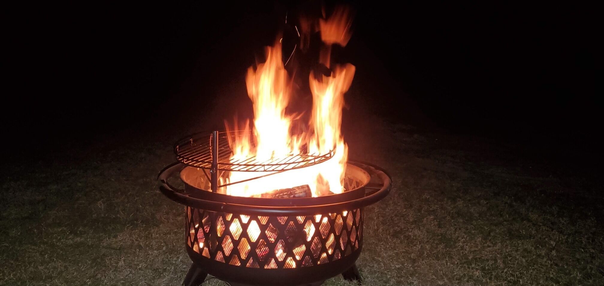 A metal fire pit with a wire grate, surrounded by a dark, grassy area.