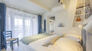 A hotel room with a bed, a chair, a window with curtains, and a shelf with decorative items. - Cavalaire sur mer Mas Boubanette (CAVALAIRE SUR MER)
