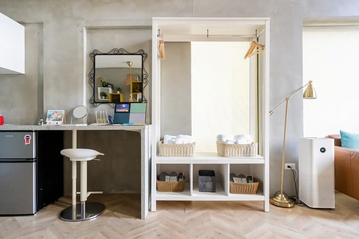 Una cocina compacta con un refrigerador, un taburete blanco, una estantería blanca con cestas y una lámpara de pie.