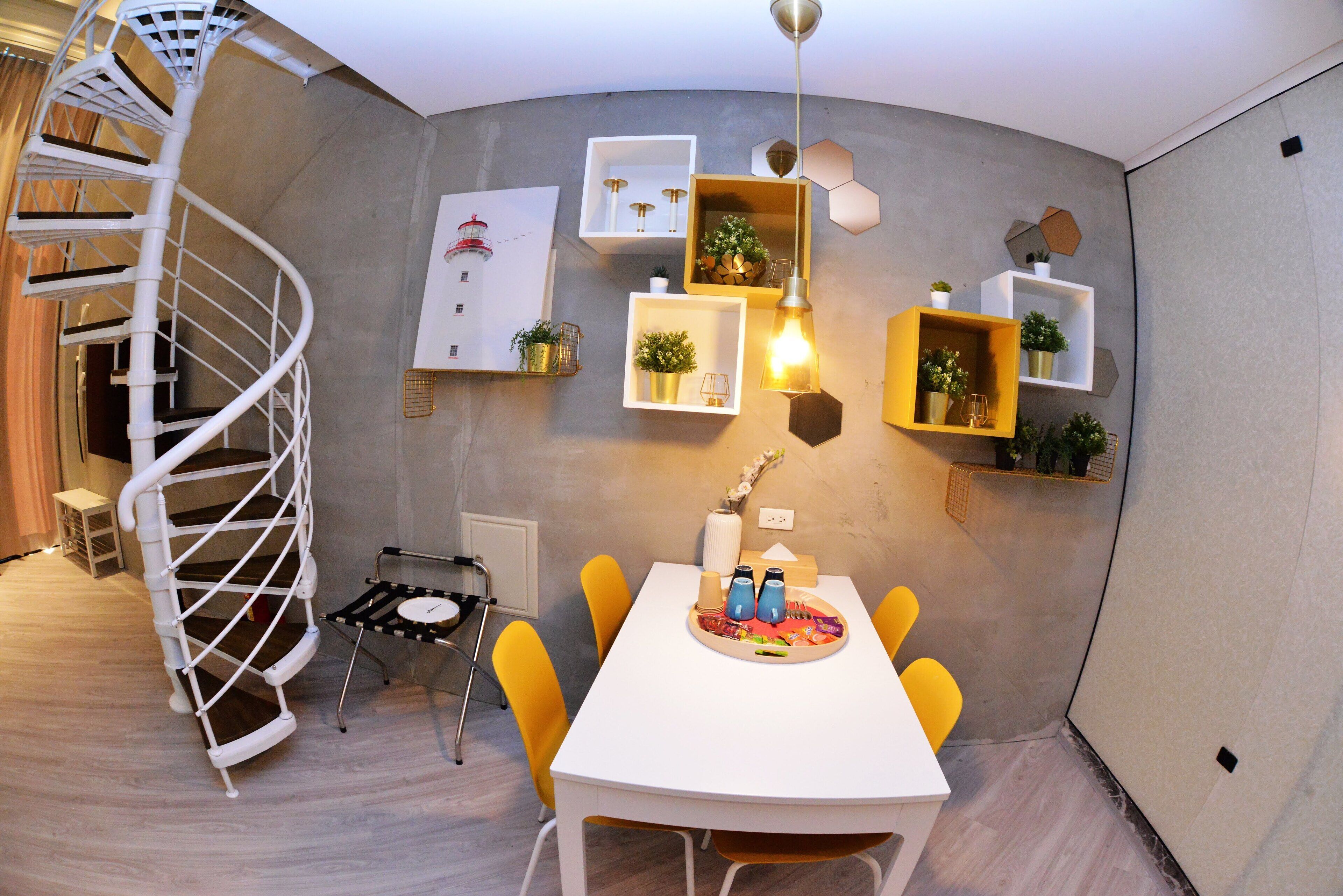 Un intérieur moderne avec un escalier en colimaçon, une table à manger entourée de chaises jaunes et divers étagères murales ornées de plantes et d’objets décoratifs.