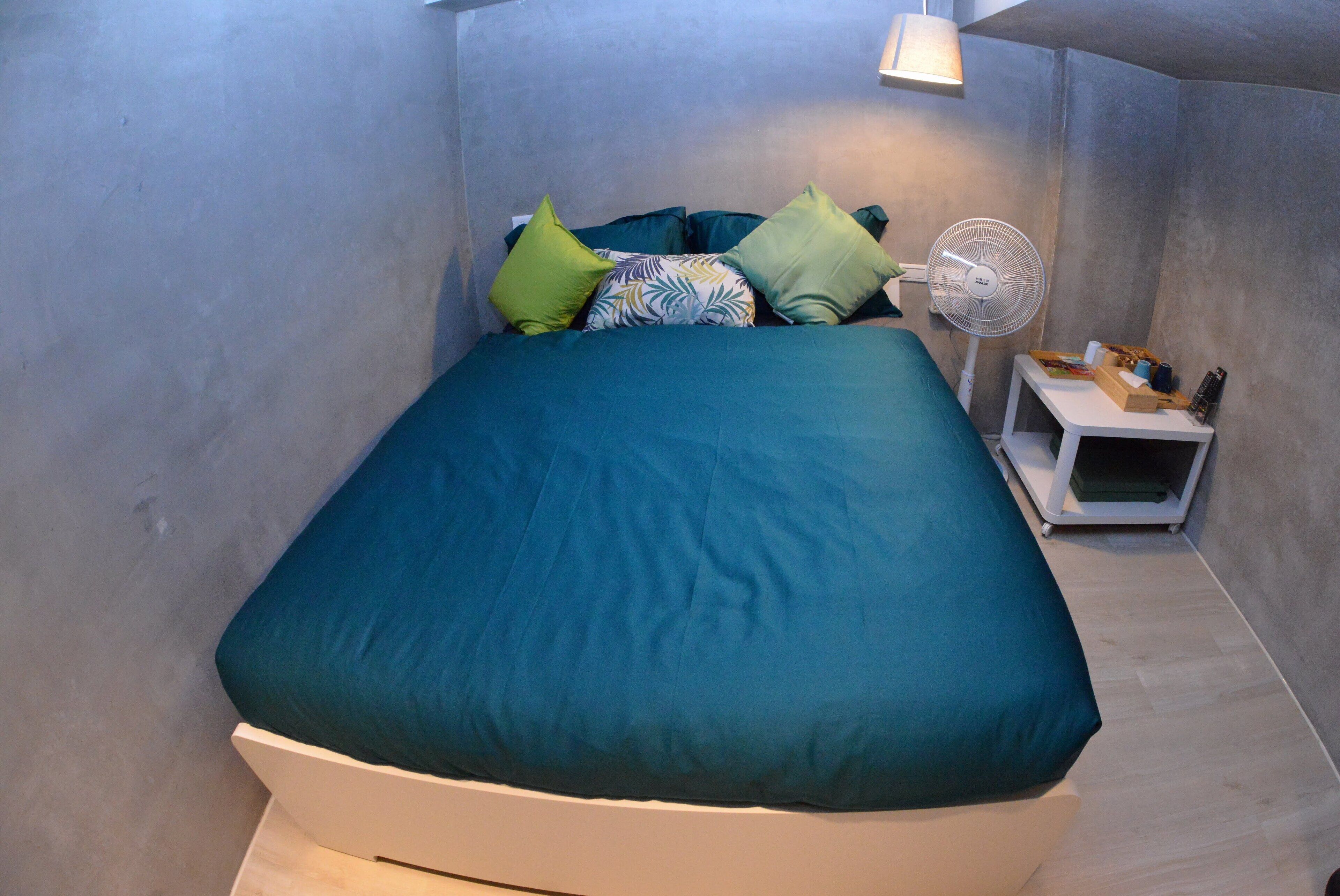 A bed with a teal bedspread and pillows, a bedside table with a fan, and a small pet bed.